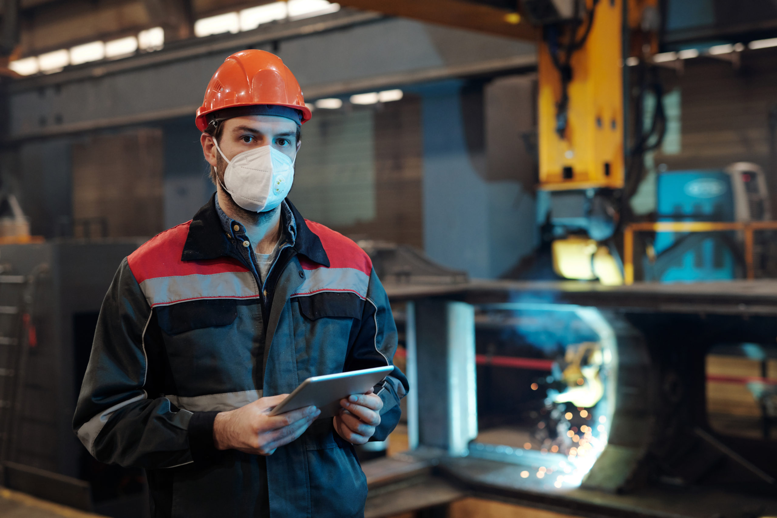 Contemporary factory engineer in protective workwear holding digital tablet while networking by his workplace in machinery plant
